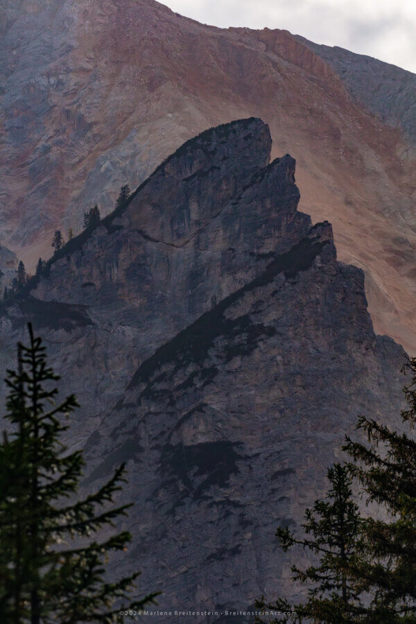 Photograph of a broken cliff, almost pyramid shaped, before an even larger mountain. The cliff looks almost like a flat wall with a ragged edge, and is streaked in blue-violet stains. A few trees adorn its angled edge. The mountain behind is bathed in warm and cool colors.