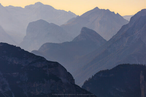 Photograph of several layers of mountains in shades of blue, against a soft yellow sky.