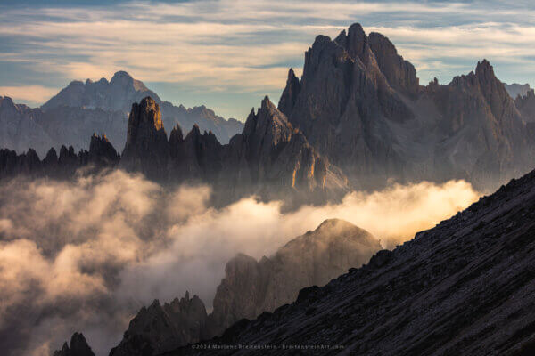 Photographs of dark and craggy mountainous spires under a clouded sky, wrapped in clouds lit by the late afternoon sun. A dark slope cuts the foreground.