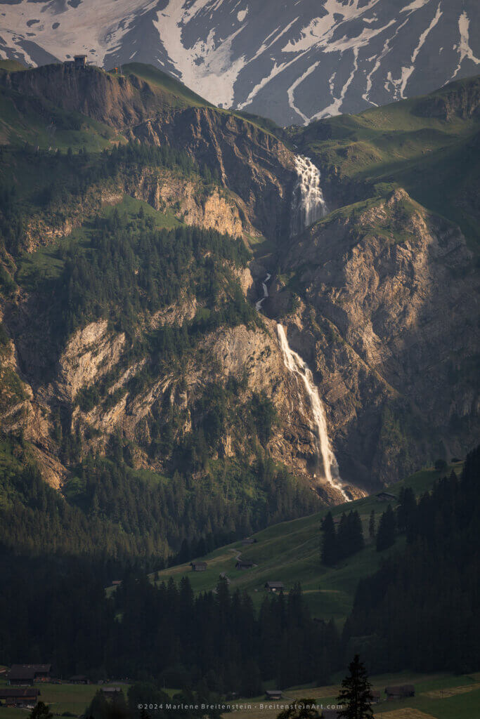 This photograph shows a green, rural, shadowed valley. It is dotted with occasional farm buildings and fields, Swiss chalets, and groves of very large spruce trees. Rising from the mid-ground is a large and rocky mountain cliff-face of tan-gray rock, with diagonal stripes, some grassy expanses, and more spruce. Its summit is topped by a tiny-looking cable car station on the left. The cliff is illuminated by a large patch of evening sunlight. From a dip in the crest appears a huge waterfall, cascading down the mountain in several steps, as it winds around the craggy surface. Above the summit is the slanted plane of an even larger mountain, striped with snow.