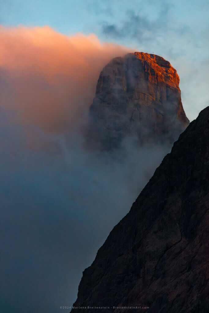 Photograph of rounded, rocky mountain peak, jutting out from behind the barren slope of another mountain. A blue-gray cloud rises between the mountains. One side of the peak is lit by the late afternoon sun. From the other side streams a dense flow of clouds, tinted a fiery orange.