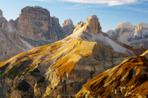 Photograph of a mountainous landscape, warmly lit by a late-afternoon sun, with a central off-white slope and peak shaped somewhat like a breast and nipple. (“Cone” doesn’t suffice. 😊) On the slopes is a rust-colored ground cover, and the mountain is backed by higher, rougher peaks.